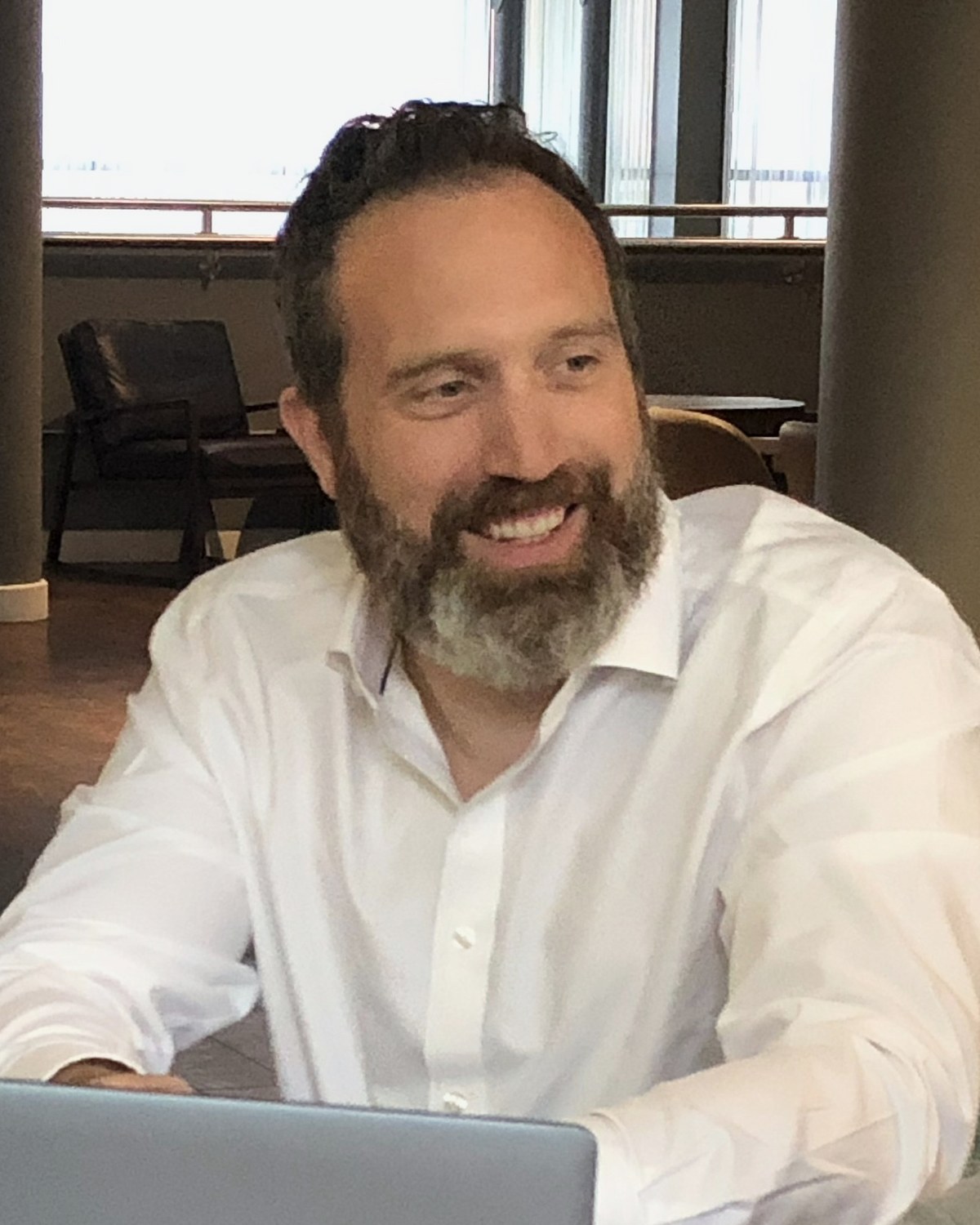 Jonathan Dee smiling at a meeting table, in a white open-neck shirt
